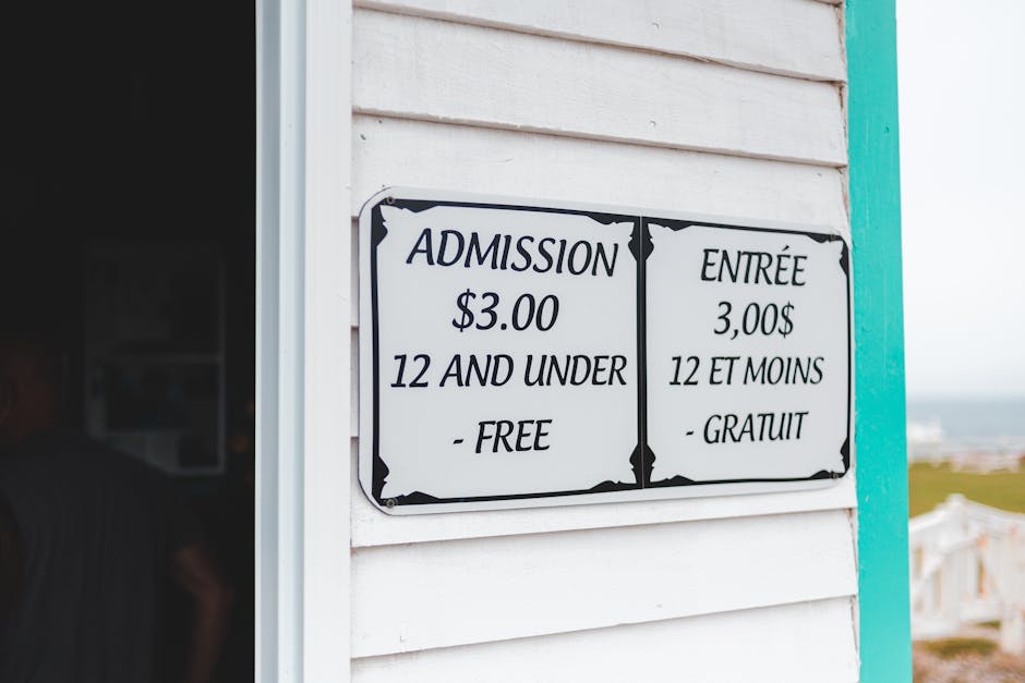 A black and white sign affixed to the exterior of a white wooden building displays admission prices in both English and French, indicating $3.00 for entry and free for children under 12 years old. The sign is mounted next to a partially open door, which reveals a dark interior space. In the background, a blurred outdoor scene suggests a seaside or park setting with distant rooftops and a cloudy sky. The building features horizontal wooden siding, and the sign's border has decorative corners. This scene may relate to entry fees for a visitor attraction, while the context of a house removal is implied by the environment, alongside the mention of moving or transportation logistics, as seen on the webpage for Man and Van Tolworth.