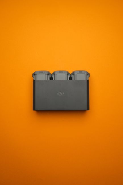 A rectangular black plastic case with a DJI logo on the front is positioned centrally against a bright orange background. On top of the case, there are three grey drone batteries with rounded edges, a