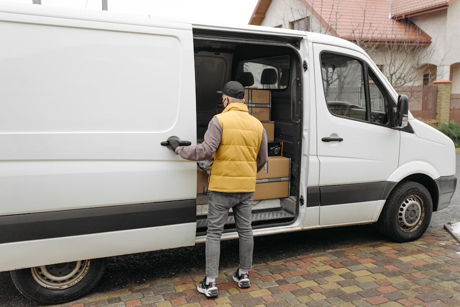 Two men are engaged in a home relocation process during daylight hours, loading a large cardboard box into the back of a white delivery van. The men are dressed casually in dark vests, long-sleeved sh