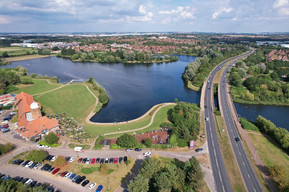 An aerial view of a residential area near Hook Rise Park, showing a large body of water with a curved shoreline surrounded by green parkland and trees. In the foreground, a building with a red tiled roof and a white tower is visible, along with a parking lot filled with various parked cars. Adjacent to the building, a pathway runs along the edge of the water, with some people walking nearby. A wide, multi-lane road with moving vehicles runs alongside the park, supported by an overpass bridge crossing the water. The background features a densely populated suburb with numerous houses and arranged streets, all set under a partly cloudy sky. The scene illustrates a peaceful neighborhood setting suitable for home relocation or furniture transport, with Man and Van Tolworth providing house removals in the area, as evidenced by their local presence and logistics expertise.