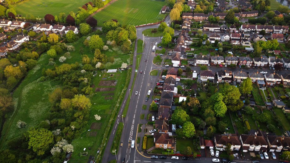 An aerial view of a residential area near Hook Rise Park, showing a large body of water with a curved shoreline surrounded by green parkland and trees. In the foreground, a building with a red tiled roof and a white tower is visible, along with a parking lot filled with various parked cars. Adjacent to the building, a pathway runs along the edge of the water, with some people walking nearby. A wide, multi-lane road with moving vehicles runs alongside the park, supported by an overpass bridge crossing the water. The background features a densely populated suburb with numerous houses and arranged streets, all set under a partly cloudy sky. The scene illustrates a peaceful neighborhood setting suitable for home relocation or furniture transport, with Man and Van Tolworth providing house removals in the area, as evidenced by their local presence and logistics expertise.