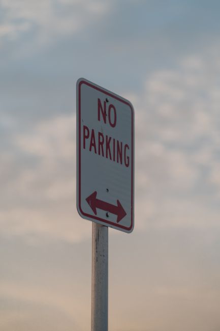 A white rectangular no parking sign with red border and red text stating 'NO PARKING' is mounted on a metal pole. Below the text, there is a black double-headed arrow indicating the restriction applies in both directions. The sign is positioned outdoors against a cloudy sky background during daylight. In the context of house removals and furniture transport, this sign may be relevant for identifying parking restrictions affecting the loading or unloading area outside a property in Tolworth. Man and Van Tolworth, a removals service provider, would need to consider such parking rules when conducting home relocation or furniture transport operations to ensure compliance and efficient access to residential premises.