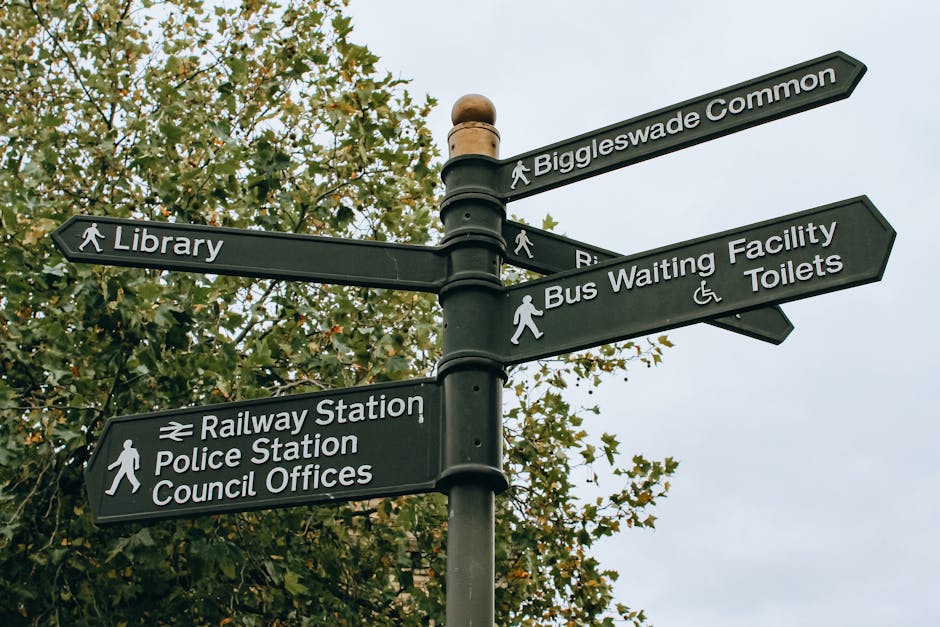 A black directional signage post with six signs pointing in different directions, located outdoors against a backdrop of green leafy trees. The signs indicate directions to Biggleswade Common, Library, Railway Station, Police Station, Council Offices, Bus Waiting Facility, and Toilets. The signs have white text and icons, with some including walking figures or accessibility symbols. The sky in the background is cloudy. This signage is situated near a pavement area, potentially within a residential or town centre environment, relevant for homeowners or movers planning walking routes or local services, such as house removals or furniture transport in Kingston or Tolworth. The description reflects the typical setting where home relocation or moving logistics are coordinated, with a focus on navigation and local amenities.