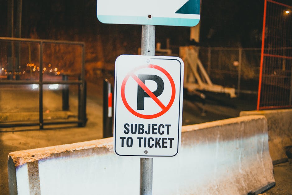 A white rectangular no parking sign with red border and red text stating 'NO PARKING' is mounted on a metal pole. Below the text, there is a black double-headed arrow indicating the restriction applies in both directions. The sign is positioned outdoors against a cloudy sky background during daylight. In the context of house removals and furniture transport, this sign may be relevant for identifying parking restrictions affecting the loading or unloading area outside a property in Tolworth. Man and Van Tolworth, a removals service provider, would need to consider such parking rules when conducting home relocation or furniture transport operations to ensure compliance and efficient access to residential premises.