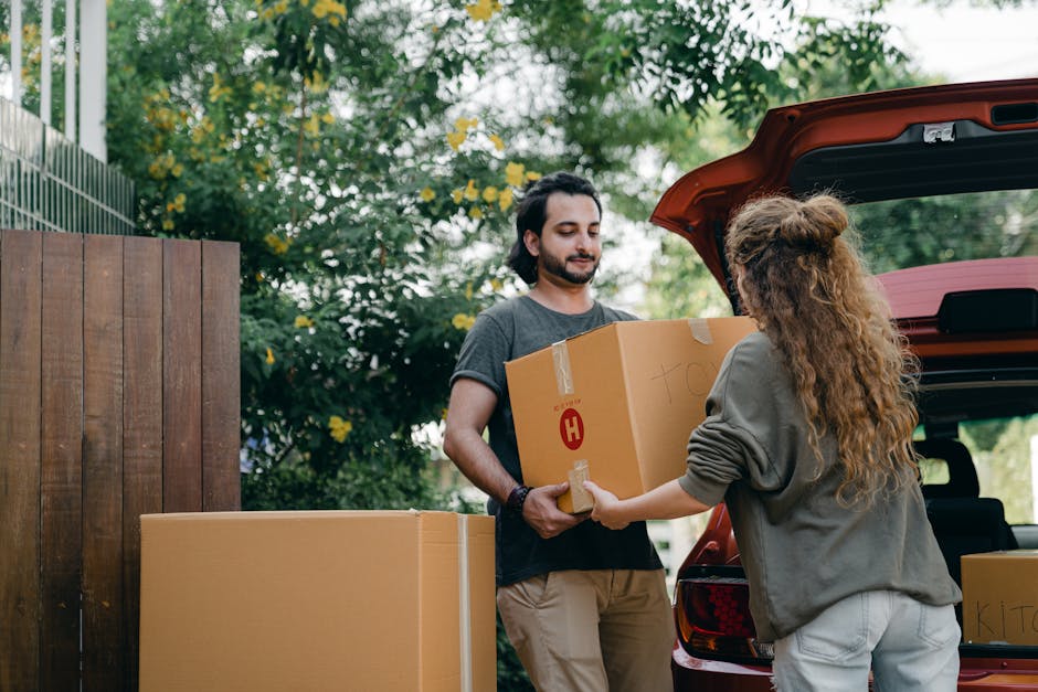 A man with dark hair, wearing a grey t-shirt and beige trousers, is standing outside near a wooden fence and green foliage, holding a cardboard box sealed with packing tape and marked with the letter 'H'. A woman with long, curly hair, dressed in a grey hoodie and light-colored trousers, is handing the box to him from the open trunk of a red vehicle, which appears to be a van or estate car, parked on a paved area. Several other cardboard boxes, some larger and some smaller, are arranged nearby on the ground and appear to be part of a home relocation or furniture transport process. The scene is lit by natural daylight, suggesting it is daytime, and captures a moment during packing or loading for a house move, illustrating a typical step in the packing and moving logistics managed by [COMPANY_NAME].
