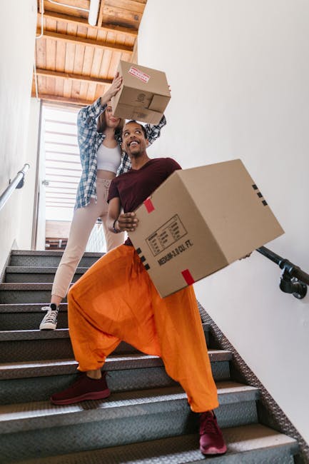 Two movers from Man and Van Tolworth are carrying cardboard boxes down a staircase inside a residential property. One mover, wearing bright orange trousers and maroon shoes, is holding a large box with labels, while the other, dressed in casual beige trousers and a plaid shirt, is passing a smaller box to their colleague. The staircase has carpeted steps and a wooden wall with paneling and a window, allowing natural light to illuminate the scene. Moving equipment such as a black hand truck or trolley is visible in the background, indicating a professional home relocation process. The image depicts the loading or unloading phase of furniture transport and packing during a same-day house removal in Tolworth, as part of the services provided by Man and Van Tolworth. This scene illustrates the careful handling and transfer of boxes typical in home removals to address last-minute moving delays.