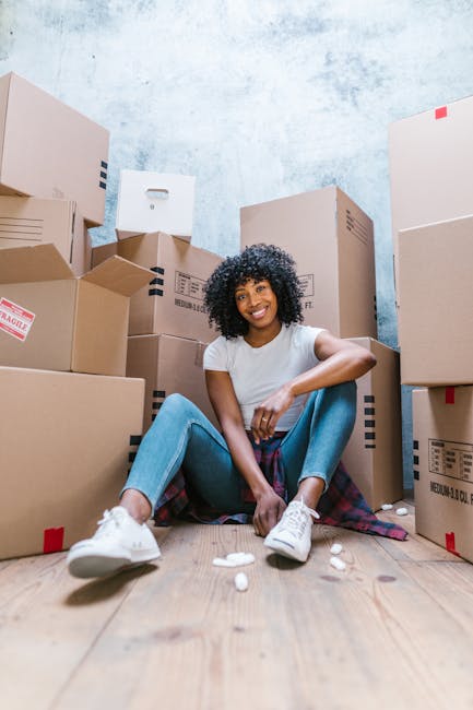 A young woman with dark curly hair and a bright smile is sitting on a wooden floor amidst a collection of packed brown cardboard moving boxes of various sizes, some labeled with 'fragile' and others with barcodes. She is dressed in a white t-shirt, light blue jeans, and white sneakers, positioned in a relaxed pose with legs apart and arms resting on her knees. Scattered on the floor are packing materials such as foam peanuts, and she is surrounded by the boxes inside a room with a light blue textured wall. The scene captures the process of home relocation, with the woman appearing ready for furniture transport and comprehensive packing and moving services provided by Man and Van Tolworth, supporting residential moves and local removals in the KT6 area.