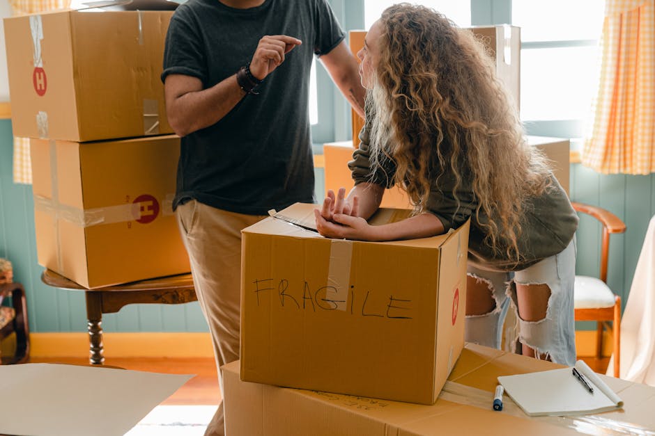 A young woman sitting cross-legged on a wooden floor surrounded by unpacked moving boxes of varying sizes in a room with textured light blue walls. She is wearing a white tank top and beige trousers, smiling as she throws packing foam into the air. An open cardboard box with a red 'Fragile' label rests on her lap, and several foam pieces are scattered around her. Behind her, there are additional stacked boxes, some sealed with red tape, and a small white shelf with a plastic bottle on it. To the right, a partially visible potted plant adds a touch of greenery. The scene captures the process of home relocation, with furniture and packing materials indicating a furniture transport and packing and moving operation. Man and Van Tolworth provide the logistical support for such house removals, exemplified by the packing and loading activities occurring inside this residence.