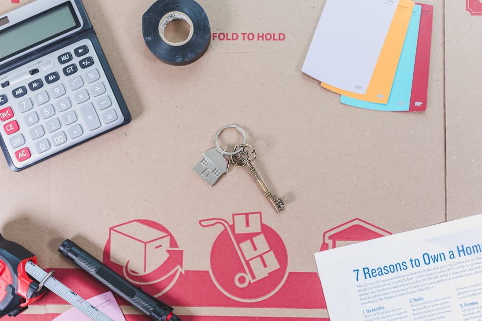 A close-up view of packing and moving supplies arranged on a flat surface, including a calculator, a roll of black tape, a set of colourful index cards, a metal house-shaped keyring with keys and a small house charm, a black marker, and a pink measuring tape, all positioned around a large red moving company logo printed on a brown cardboard packing box. In the background, there is a document titled '7 Reasons to Own a Home' partially visible, suggesting preparation for house removal or relocation. The scene is illuminated with natural light, reflecting careful organization of packing materials and tools used during furniture transport and home relocation processes, with the presence of the 'FOLD TO HOLD' instruction on the packing box indicating proper packaging methods.