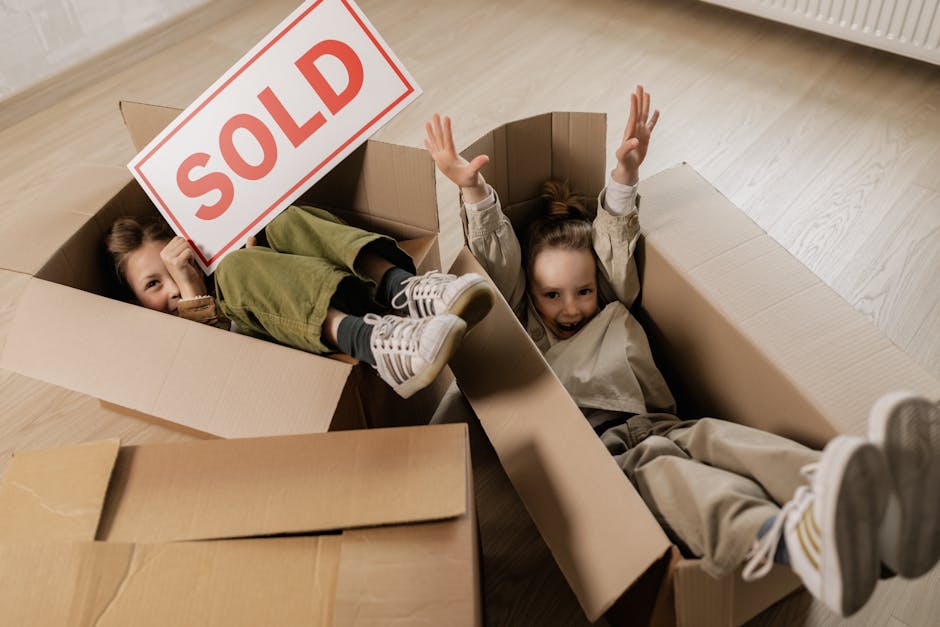 Two young children are inside large cardboard moving boxes on a light-colored wooden floor. One child, positioned further inside the box, is holding a sign that reads 'SOLD' and has their legs crossed, wearing a green outfit and white sneakers. The other child, lying on their back with arms raised, appears to be smiling and is dressed in a beige jacket and pants with white sneakers. The scene suggests a playful moment during a home relocation process, with the boxes arranged close to a wall and a radiator visible in the background. This image illustrates packing and moving activities, which are typical components of furniture transport and home removals, as handled by companies like Man and Van Tolworth.