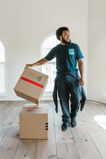 A close-up view of packing and moving supplies arranged on a flat surface, including a calculator, a roll of black tape, a set of colourful index cards, a metal house-shaped keyring with keys and a small house charm, a black marker, and a pink measuring tape, all positioned around a large red moving company logo printed on a brown cardboard packing box. In the background, there is a document titled '7 Reasons to Own a Home' partially visible, suggesting preparation for house removal or relocation. The scene is illuminated with natural light, reflecting careful organization of packing materials and tools used during furniture transport and home relocation processes, with the presence of the 'FOLD TO HOLD' instruction on the packing box indicating proper packaging methods.