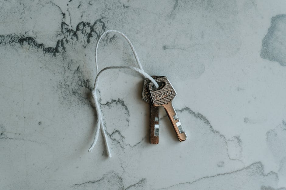A close-up image of two metal keys, one with a rectangular head and the other with a round head, attached together by a white string. The keys are resting on a light grey, marbled surface that may be a countertop or tiled floor. The keys are unused and appear clean, with visible grooves for lock mechanisms. This image represents the initial access or key handover stage during house removals or home relocation, often facilitated by professional moving companies such as Man and Van Tolworth. It highlights the importance of secure key transfer in the process of packing, loading, and transporting household items, including furniture and boxes, during a move. The lighting is natural and even, emphasizing the metallic texture and simple composition related to moving logistics and access preparation, aligned with the topic of Tolworth Broadway house access tips for house removals.