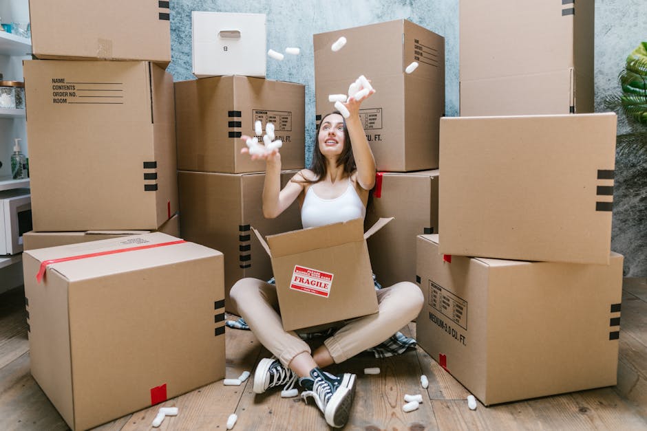 A young woman with long dark hair, sitting cross-legged on a wooden floor inside a room, surrounded by several large cardboard boxes, some with visible packing labels and red 'Fragile' stickers. She is wearing a white sleeveless top, beige pants, and black and white sneakers. The woman is mid-action, throwing white foam packing peanuts into the air with both hands, with a cheerful expression on her face. The boxes are stacked on either side and behind her, and some are partially open, revealing additional packing materials inside. To the left, there is a small countertop with a potted plant and a microwave, indicating a domestic environment. The scene is well-lit, suggesting daytime, capturing the process of packing or unpacking during a home relocation or furniture transport, consistent with house removal services provided by Man and Van Tolworth, as part of a furniture transport or packing and moving operation.