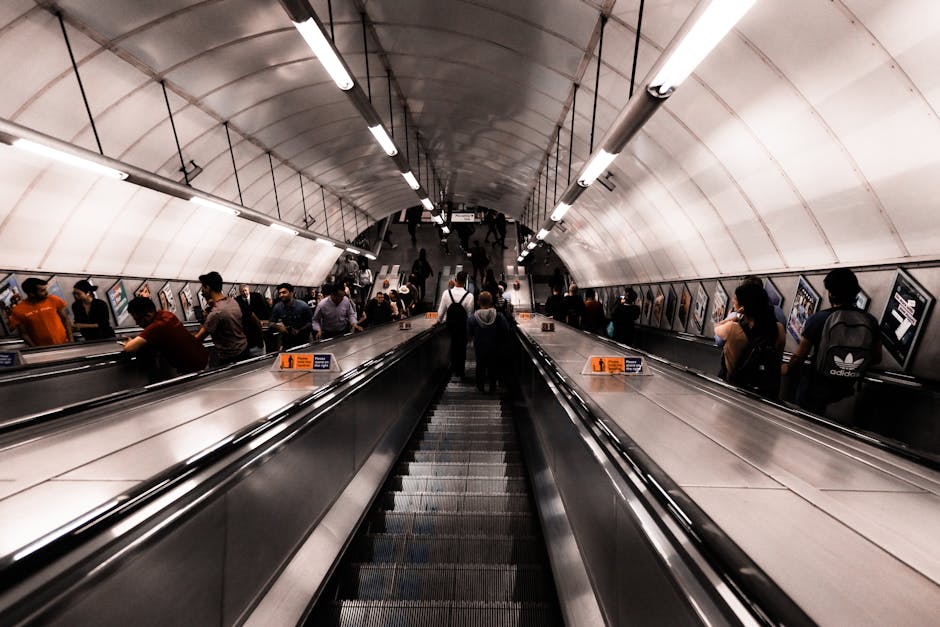 Inside a busy underground station with a curved, tunnel-like ceiling illuminated by long fluorescent lights. An escalator is centrally positioned, with people riding up and down while others walk along the platform and stairs at the top. The scene captures the movement of individuals, some carrying bags or personal belongings, amidst advertisements mounted along the side walls. The environment has a modern, utilitarian design typical of urban transport hubs, with clean metal and concrete surfaces. This setting relates to the context of home relocation or moving logistics, as part of the process involving travel and transport to or from residential premises, consistent with removal services offered by Man and Van Tolworth.