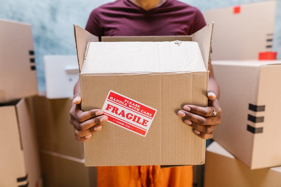 A person standing inside a room, holding an open cardboard box with both hands, displaying a red and white 'Fragile Handle with Care' sticker on the front. The individual is wearing a maroon top, and their hands are visible, one of which has a ring on the ring finger. Several other closed cardboard boxes, some with black tape, are stacked around them on the floor, suggesting a packing or home relocation process. In the background, there are blurred elements indicating an indoor environment, with a light wall and some additional boxes, supporting the scene of furniture transport and packing during a house move, as coordinated by services such as Man and Van Tolworth.