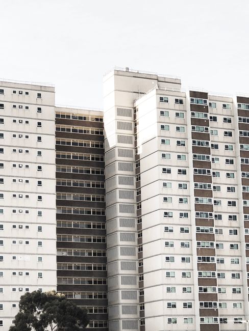 A tall, multi-storey high-rise building with a modern exterior composed of light grey and dark grey panels, featuring numerous evenly spaced rectangular windows, some of which are slightly open. The b