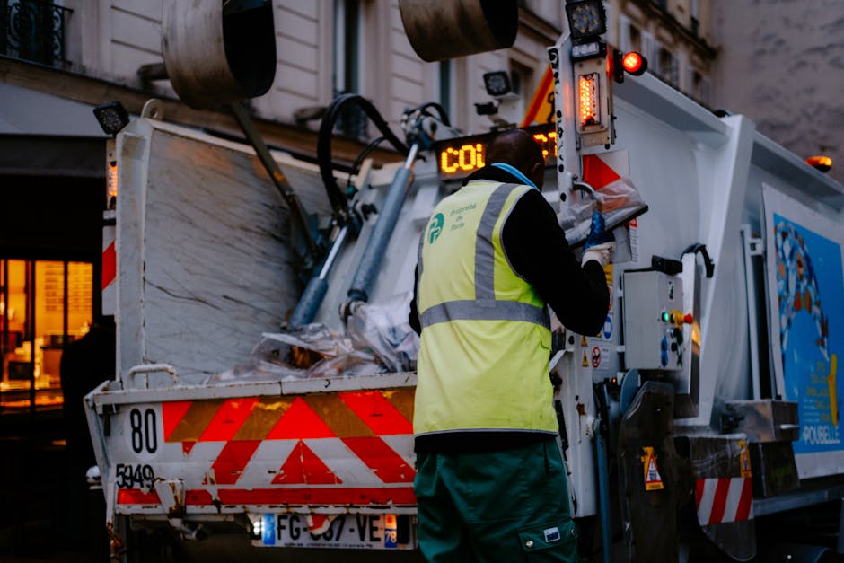 A worker from Man and Van Tolworth, dressed in a high-visibility yellow vest and dark clothing, operates a waste disposal vehicle during an urban home relocation. The worker is seen from behind, using a control panel on the side of the vehicle, which is positioned on a street or driveway. The vehicle's rear opening is filled with cardboard boxes, plastic wrapping, and packaging debris, indicating a cleanup or removal process following furniture transport and packing activities. The scene is set in low evening light, with the vehicle's red and white reflective markings visible, and a residential building with illuminated windows in the background. This image exemplifies the loading and waste disposal steps involved in house removals, highlighting the importance of proper waste management during a relocation in Tolworth, as managed by Man and Van Tolworth.