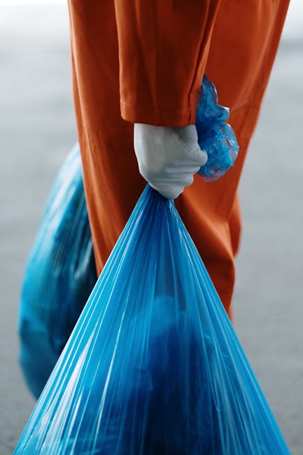 Close-up of a person wearing an orange protective suit and white gloves, holding a large blue trash bag filled with waste or discarded items, standing outdoors near a paved area. In the background, part of a vehicle or moving van with an open door can be seen, indicating a loading or unloading activity. Inside the vehicle, several cardboard boxes, some wrapped in plastic or packing material, and other household packing supplies are visible, arranged on the vehicle’s floor. The scene suggests an organized removal or waste disposal process, with items ready for transport or disposal, typical of house removals or relocation services by Man and Van Tolworth. The environment is well-lit, showcasing a professional approach to packing, loading, and waste management during home relocation activities, aligned with the topic of waste disposal rules and removals responsibility in Tolworth.
